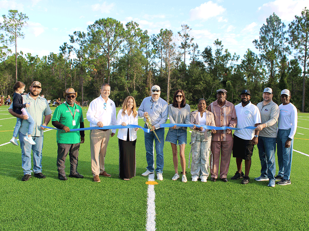 Ten other people stand behind a ribbon on a football field while the figure in the middle, District 3 Commissioner Clay Murphy, cuts the ribbon with huge golden scissors: Project Manager Chris Gatchell, West Augustine Community Redevelopment Agency Steering Committee Chair Robert Nimmons, Parks and Recreation Director Ryan Kane, District 5 Commissioner Ann Taylor, District 2 Commissioner Sarah Arnold, West Augustine Community Redevelopment Agency Steering Committee Co-Chair Dwala Willis, West Augustine Historical Community Development Corporation Founder and President Greg White, Public Works Director Greg Caldwell, and two other members of the West Augustine community
