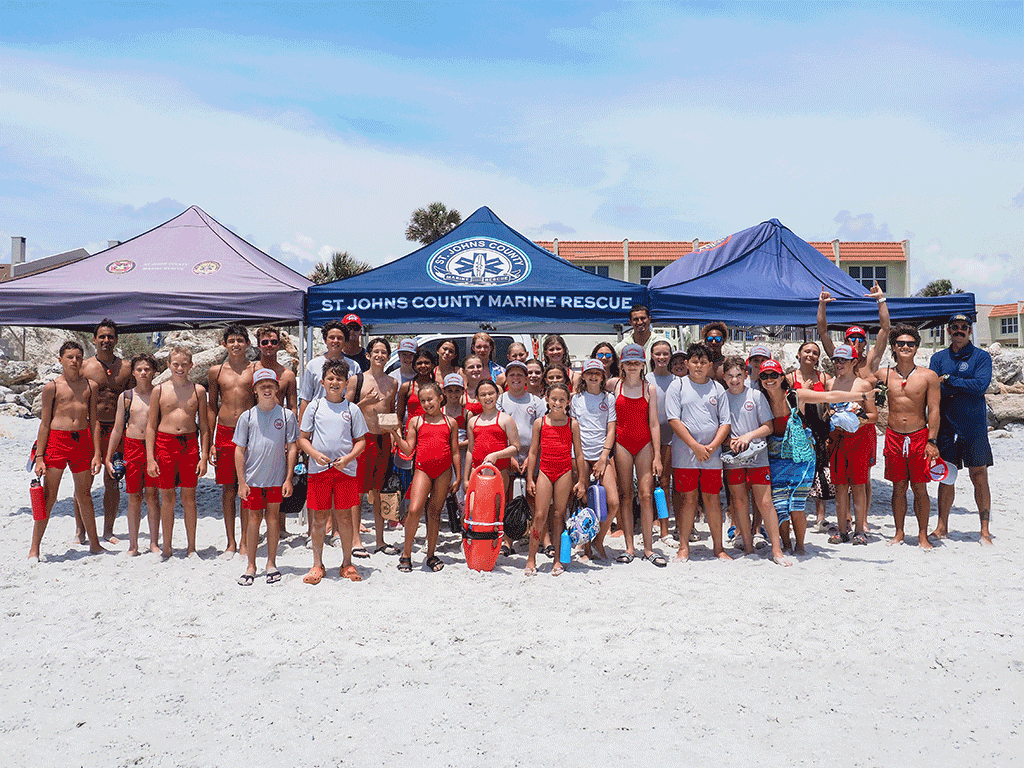 A group of kids in red swimsuits stand for a picture on the beach beneath a tent that says "St. Johns County Marine Rescue"