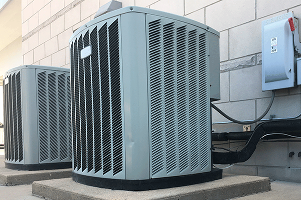 two heating/cooling units outside of a block building sitting on concrete pads. On/off power breaker switch attached to wall.