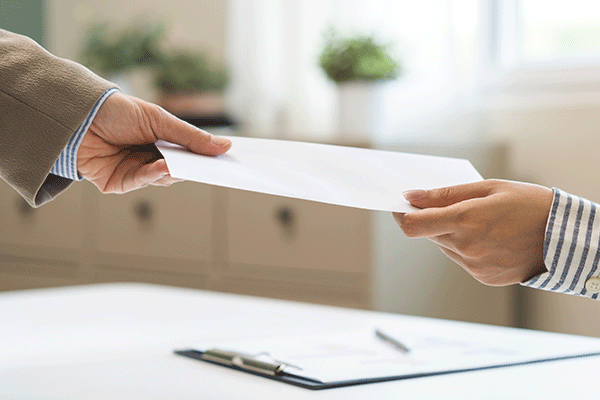 One hand passing a white business-sized envelope to someone else's hand. Both are in business attire.
