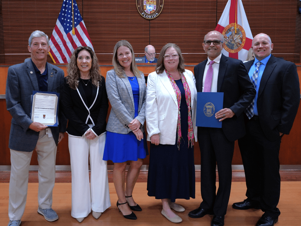 Six individuals in formal attire pose in government auditorium setting with American Flag and Florida State Flag in the background