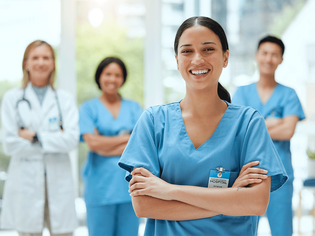Four smiling healthcare workers with arms crossed. Three are in out-of-focus background. A young dark-haired female is in the foreground.