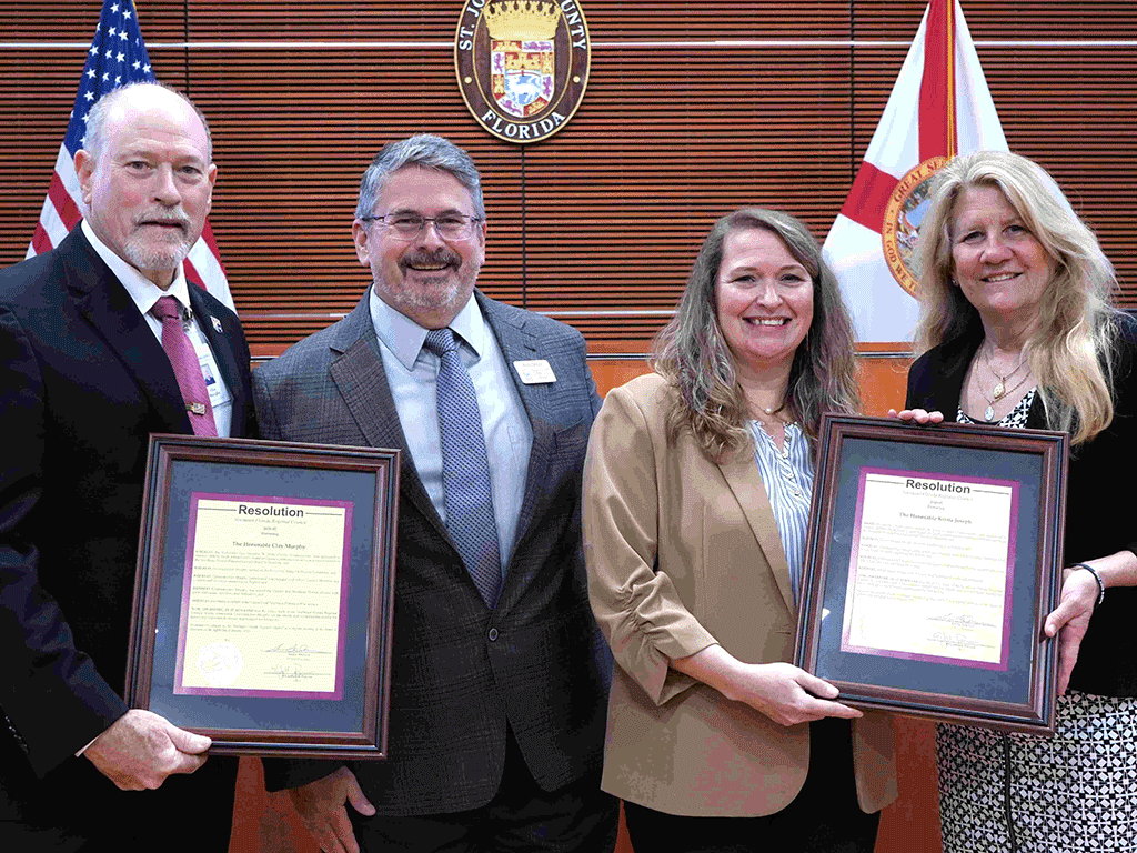 Four individuals in formal attire in an auditorium. Two holding framed certificates.