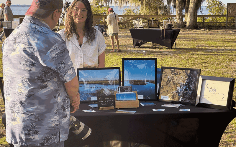 A man with a camera talks to a woman at an outdoor stall displaying her nature photography.