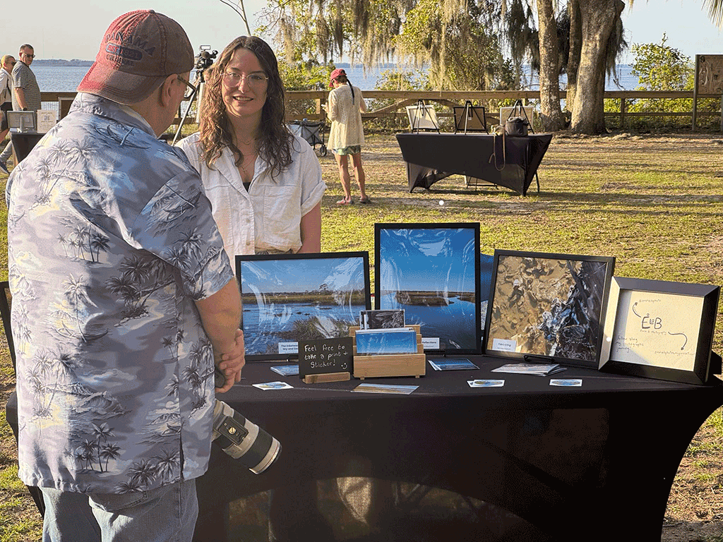 A man with a camera talks to a woman at an outdoor stall displaying her nature photography.