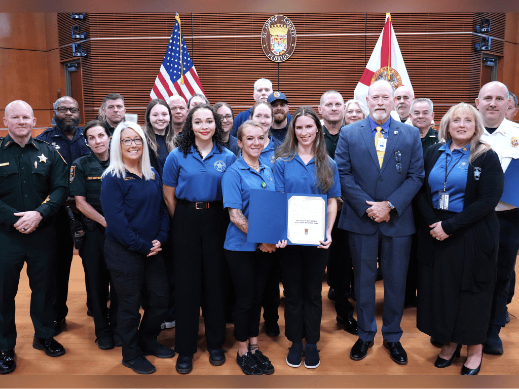 Group of fire rescue and sheriff’s office personnel pose in a government auditorium with a certificate and the American Flag, Florida State Flag and the St. Johns County seal in the background