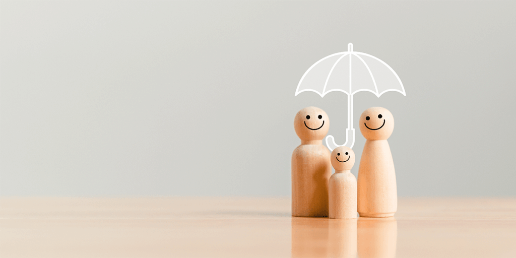 Three simplistic wooden figurines smiling and holding a transaprent umbrella above them. Neutral background and wooden tabletop surface.