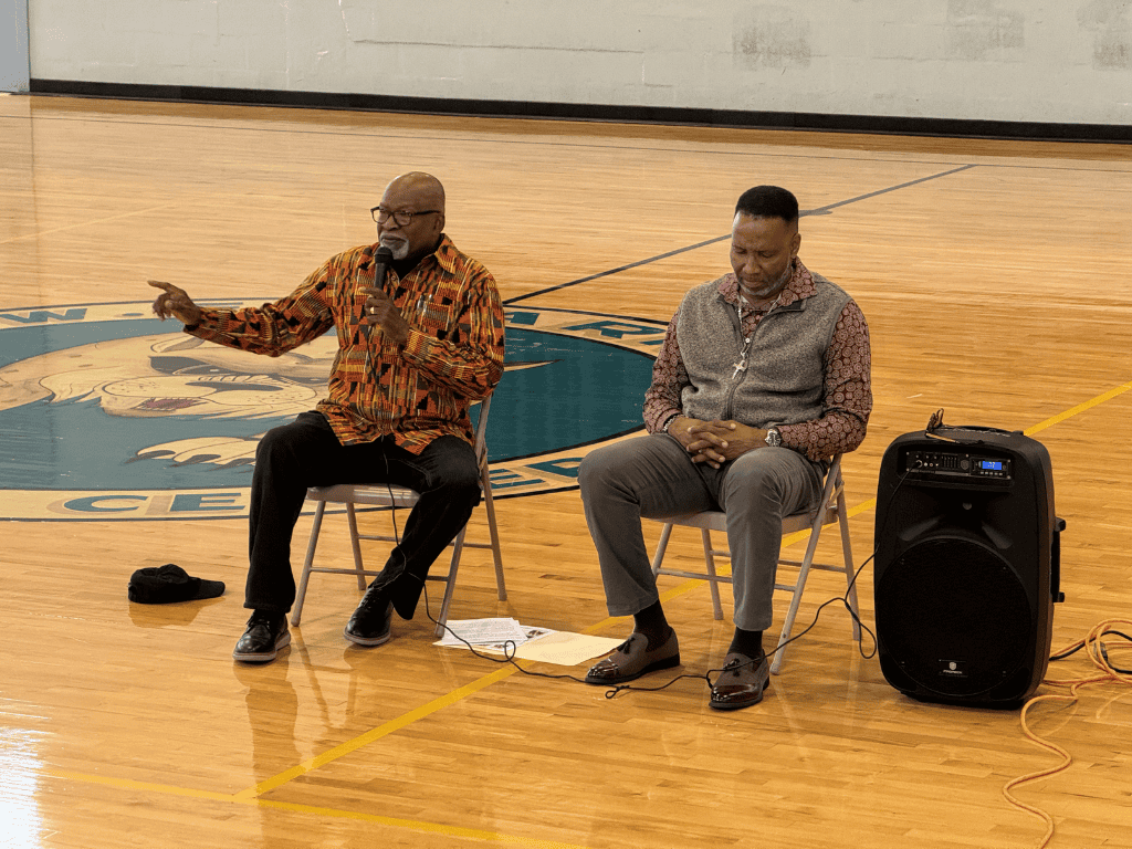 Two individuals sit on chairs placed on a gymnasium floor with a school logo visible. One is speaking while gesturing, and the other listens. There's a speaker and some papers on the floor beside them.