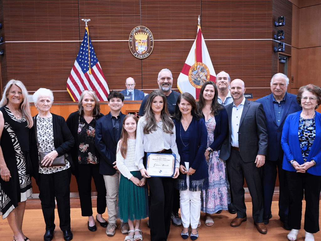 A group of individuals stand in an auditorium, one holding a certificate. The US flag, Florida State Flag, and the St. Johns County seal are in the background.