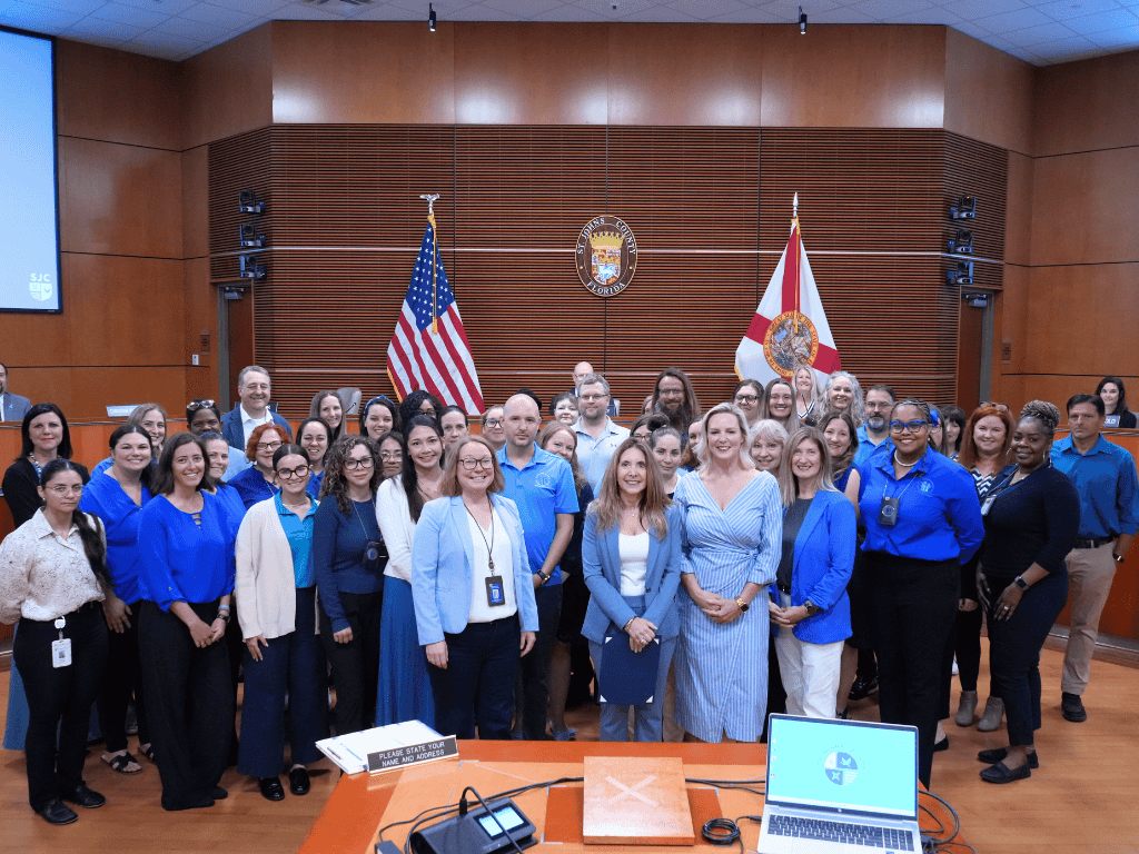 A group of individuals stand in a government auditorium, one holding a certificate, with the American Flag, Florida State Flag, and St. Johns County seal in the background.