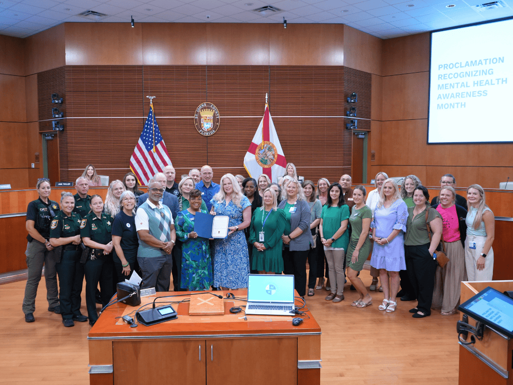 A group of individuals stand in an auditorium, one holding a certificate. The US flag, Florida State Flag, and the St. Johns County seal are in the background.