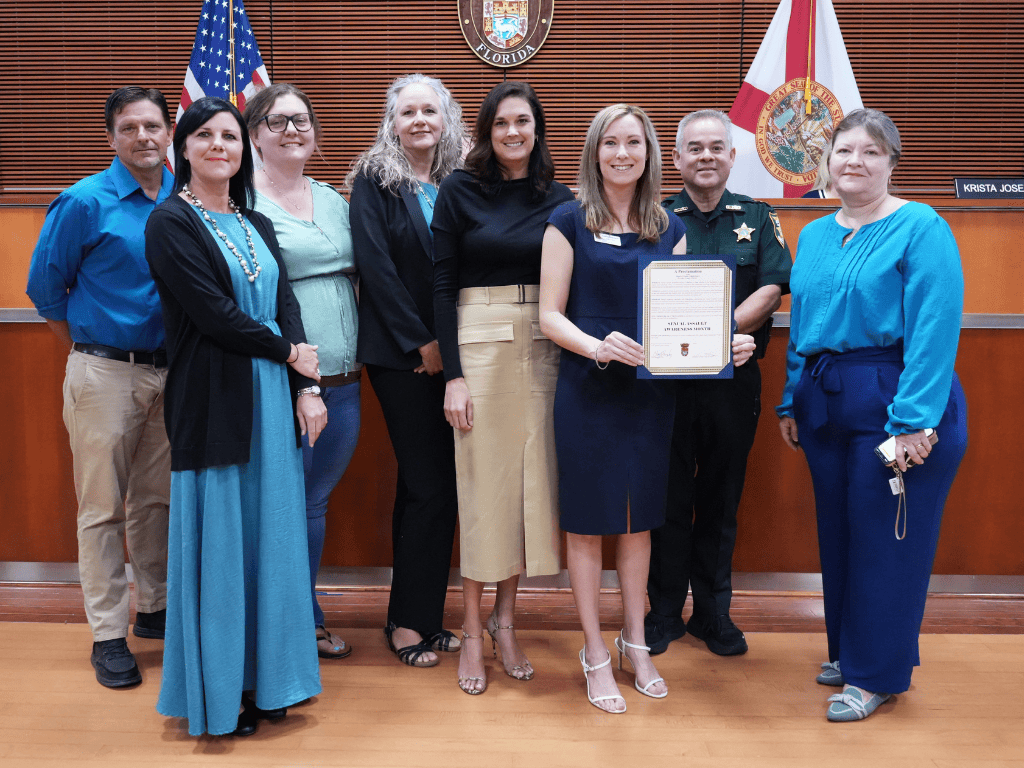 A group of individuals stand in a government auditorium, one holding a certificate, with the American Flag, Florida State Flag, and St. Johns County seal in the background.