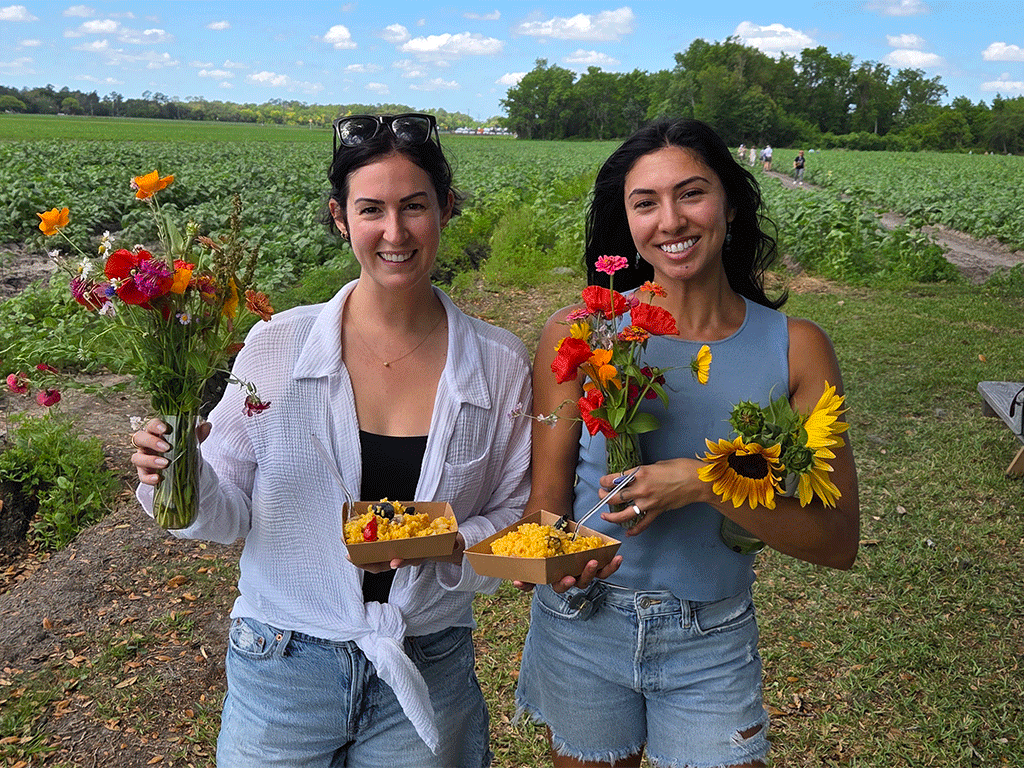 two smiling women holding vases of several fresh cut flowers and bowls of food with farm crops in the background