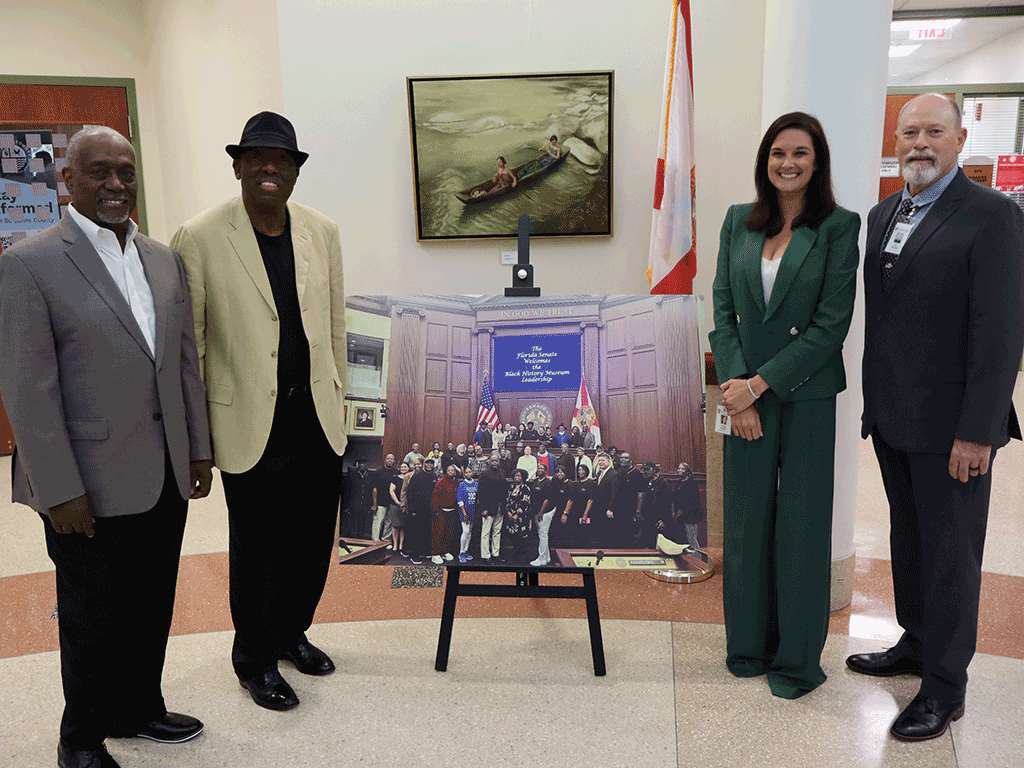 Commissioners Murphy and Arnold stand with two others in front of a photo of group of people.