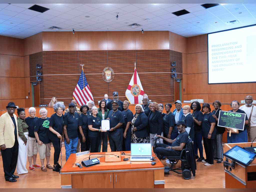 A group of individuals stand in an auditorium, one holding a certificate. The US flag, Florida State Flag, and the St. Johns County seal are in the background.