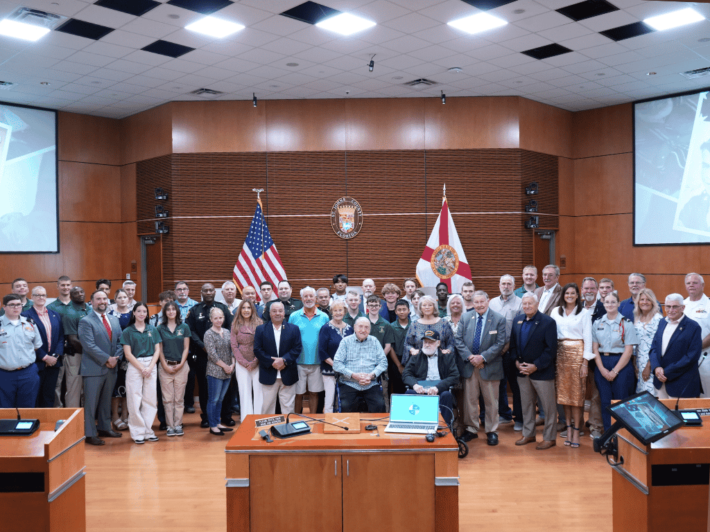 Group of individuals standing in an auditorium setting, one holding a certificate, with the American Flag, Florida State Flag, and the seal of St. Johns County in the background.