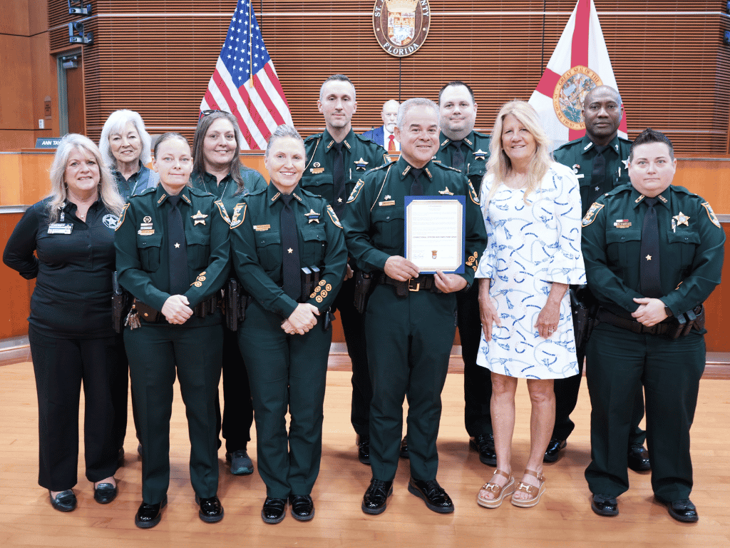 Group of individuals standing in an auditorium setting, one holding a certificate, with the American Flag, Florida State Flag, and the seal of St. Johns County in the background.