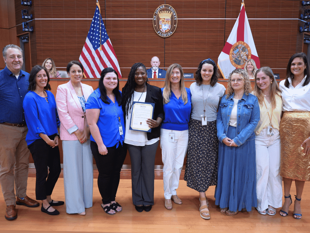 Group of individuals standing in an auditorium setting, one holding a certificate, with the American Flag, Florida State Flag, and the seal of St. Johns County in the background.