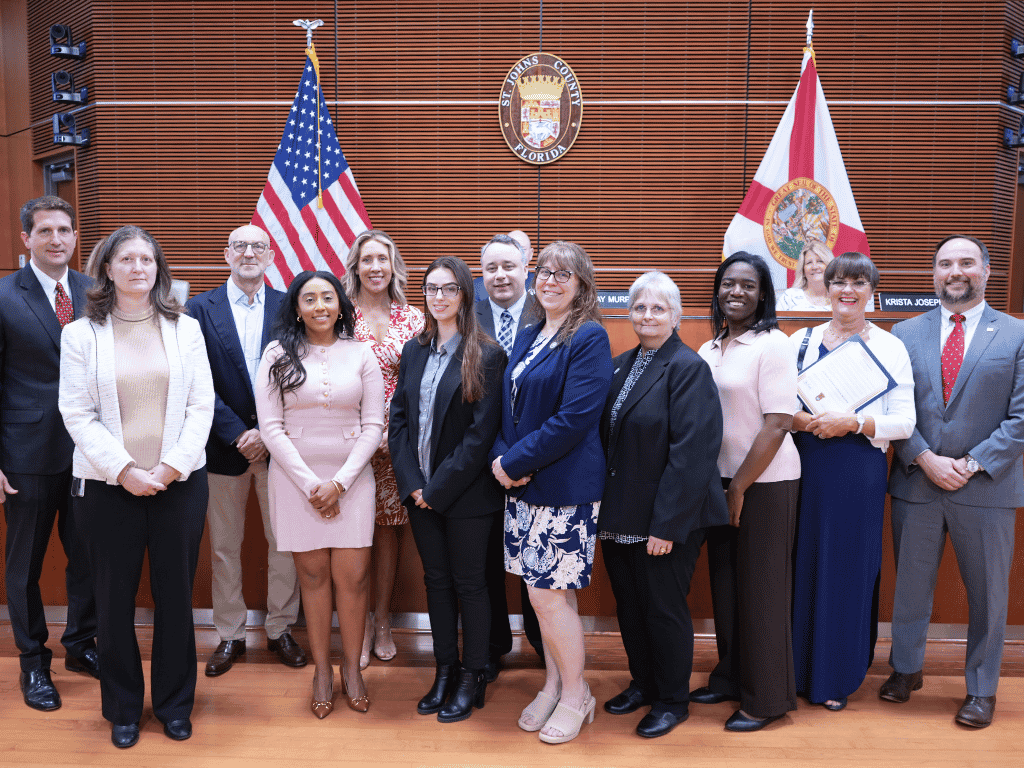 Group of individuals standing in an auditorium setting, one holding a certificate, with the American Flag, Florida State Flag, and the seal of St. Johns County in the background.