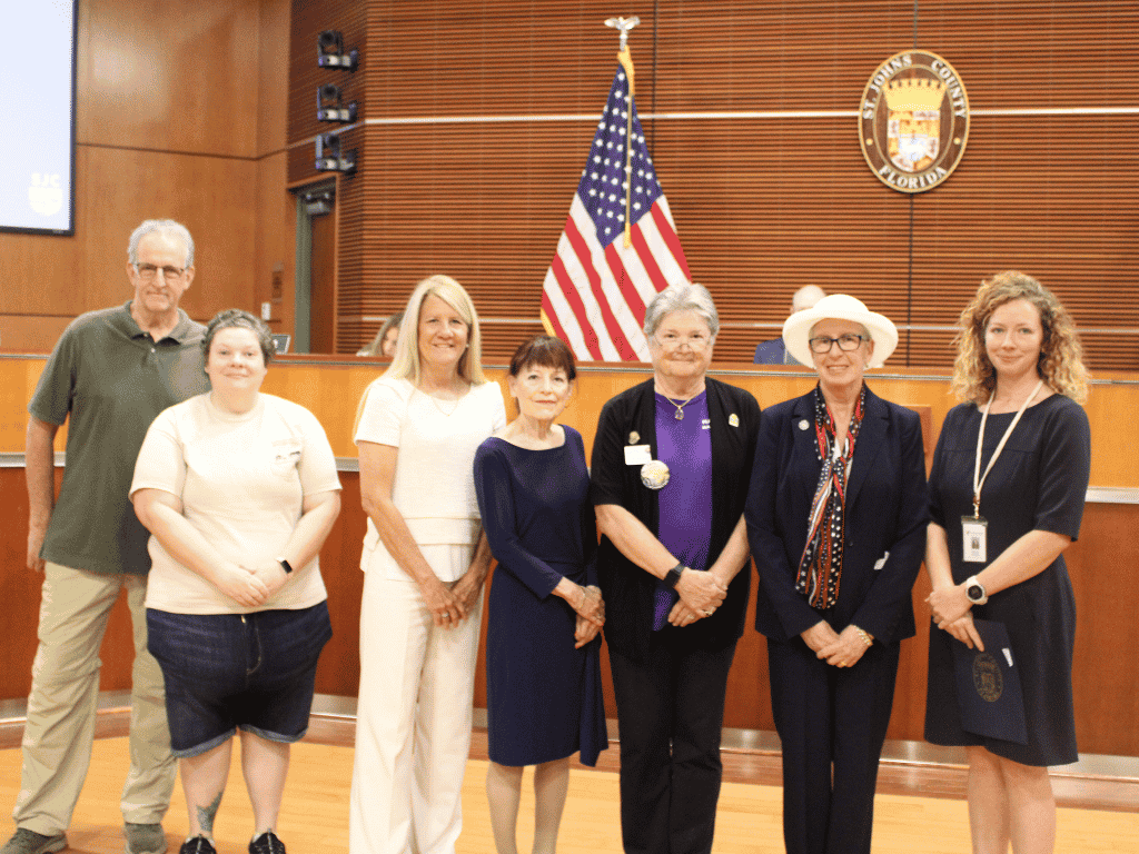 Individuals standing in an auditorium, one holding a certificate, with the St. Johns County Seal, the American Flag, and the Florida State Flag in the background.