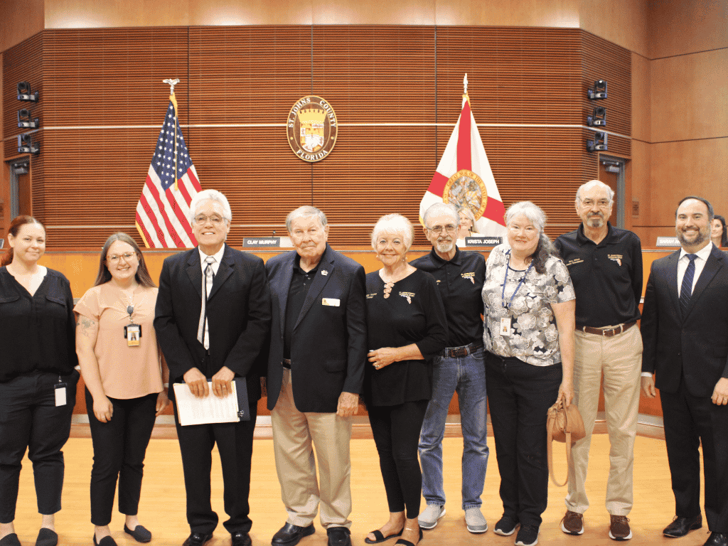 Individuals standing in an auditorium, one holding a certificate, with the St. Johns County Seal, the American Flag, and the Florida State Flag in the background.