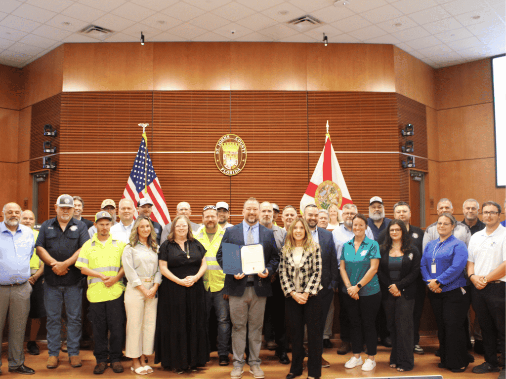 Individuals standing in an auditorium, one holding a certificate, with the St. Johns County Seal, the American Flag, and the Florida State Flag in the background.