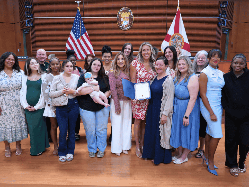 Group of individuals standing in an auditorium setting, one holding a certificate, with the American Flag, Florida State Flag, and the seal of St. Johns County in the background.