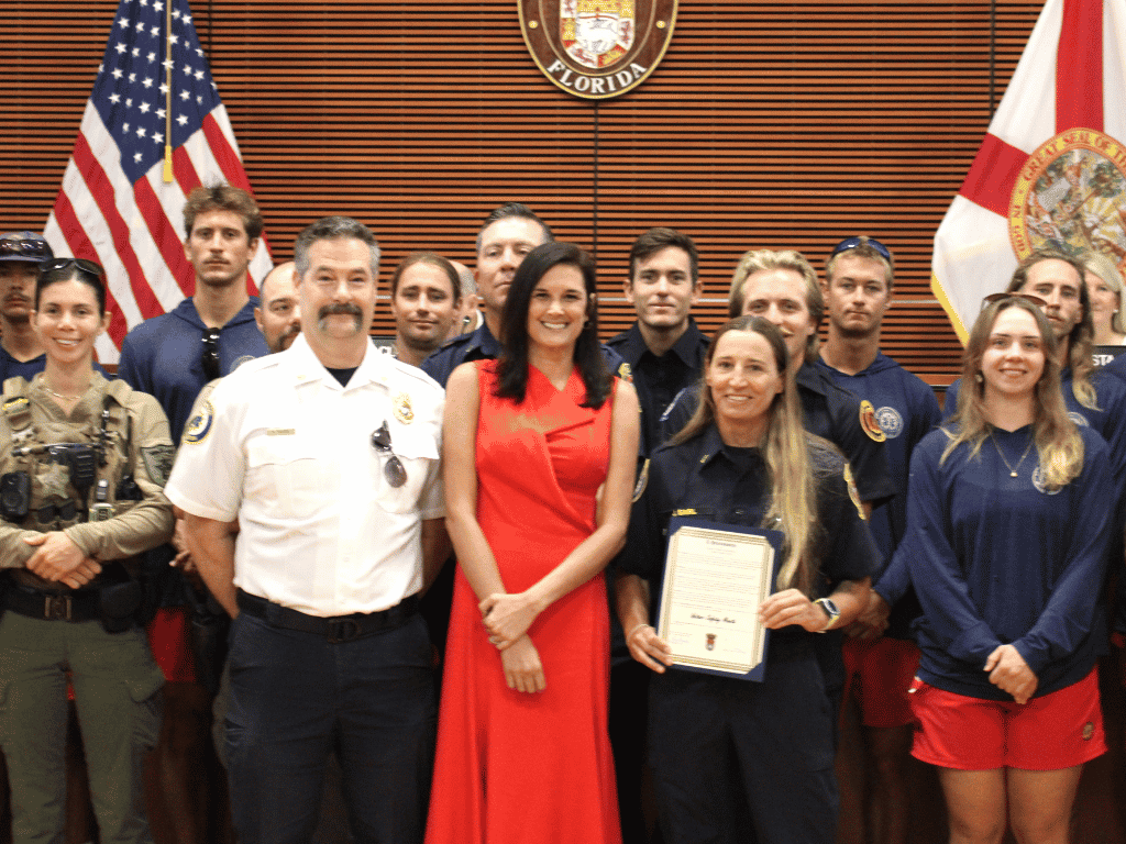 Individuals standing in an auditorium, one holding a certificate, with the St. Johns County Seal, the American Flag, and the Florida State Flag in the background.