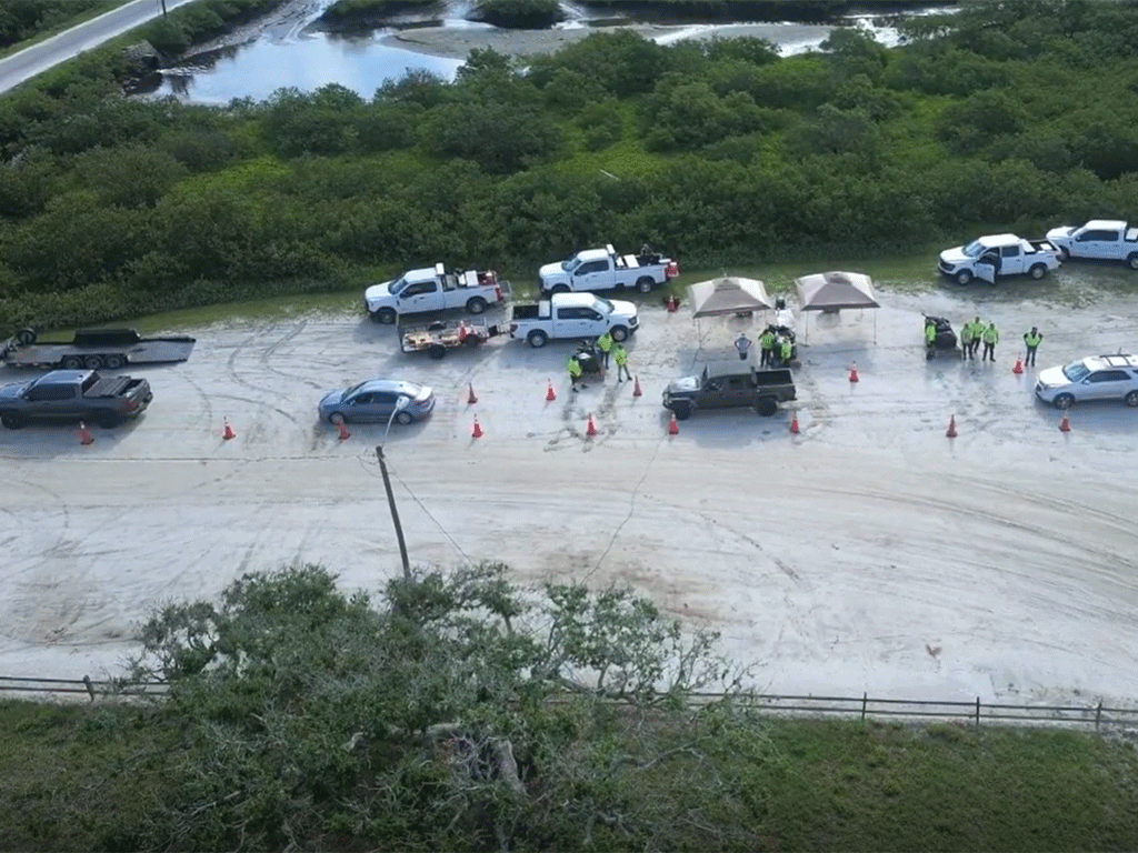 Aerial view of sandbag distribution link with cars, trucks, canopies, traffic cones, and workers wearing hi-viz attire.
