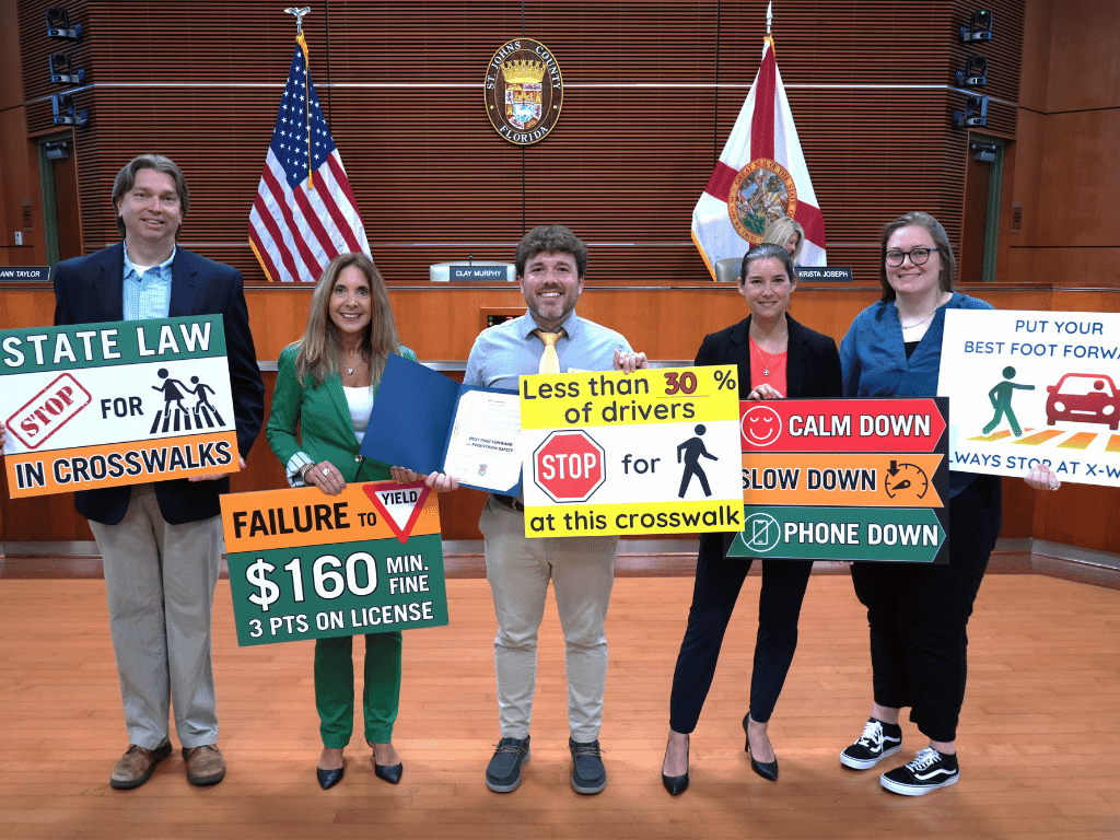 Group of individuals post in auditorium holding signs about pedestrian safety with the American flag, Florida flag, and St. Johns County Seal in the background.