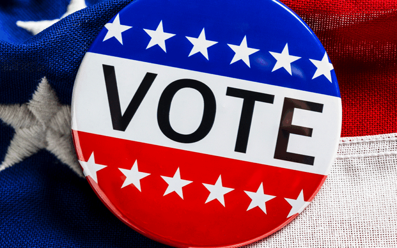 Red, white, and blue button with stars and text reading VOTE with an American flag as the background