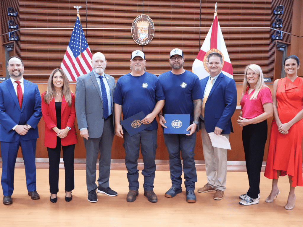 Individuals posing together. Two holding certificates. The American flag, Florida state flag, and the St. Johns County seal in the background.