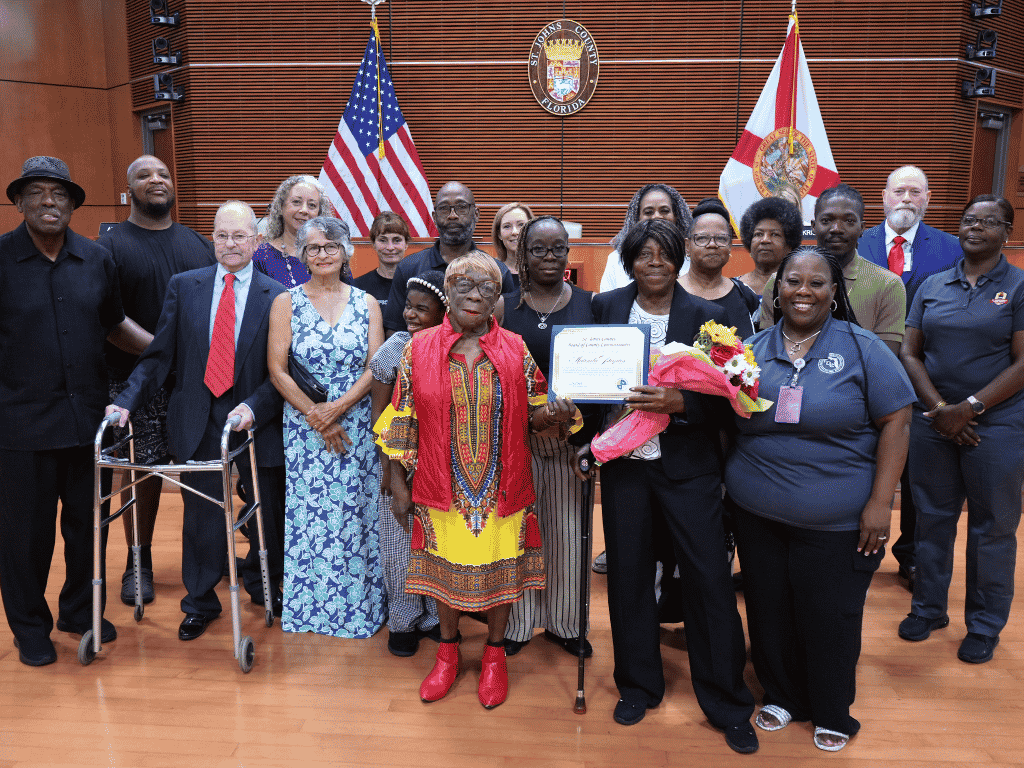 Individuals stand together, one holding a certificate, with the American Flag, Florida flag, and St. Johns County Seal in the background