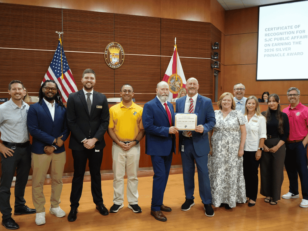 Individuals stand together, one holding a certificate, with the American Flag, Florida flag, and St. Johns County Seal in the background