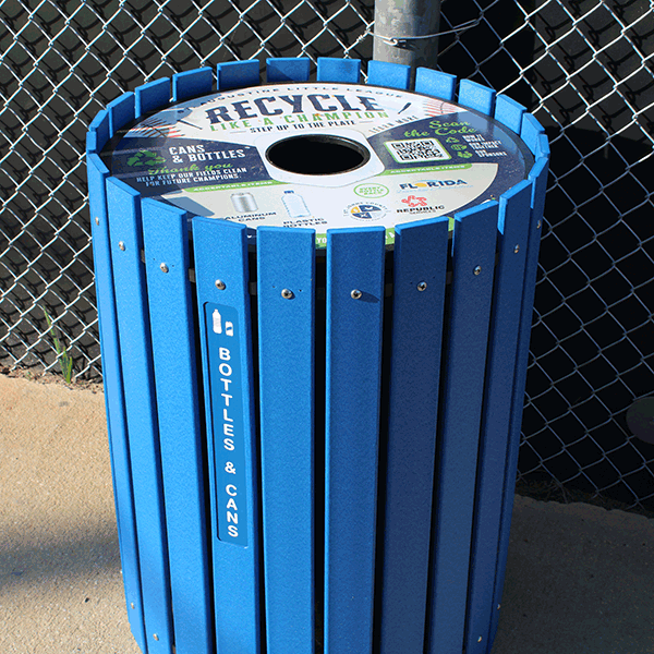 Tall blue bin constructed with wooden vertical slats used for collecting plastic and metal cans for recycling. Top cover with hole for depositing items with various text, logos, QR code and graphics.