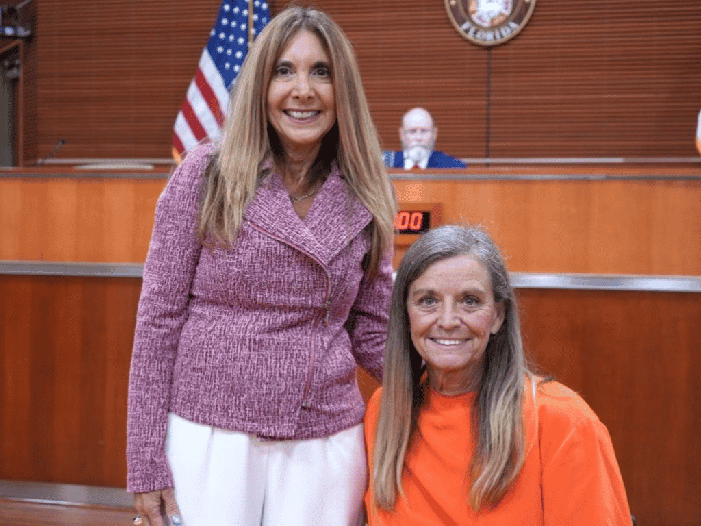 Two individuals pose together with the American Flag and the St. Johns County Seal in the background