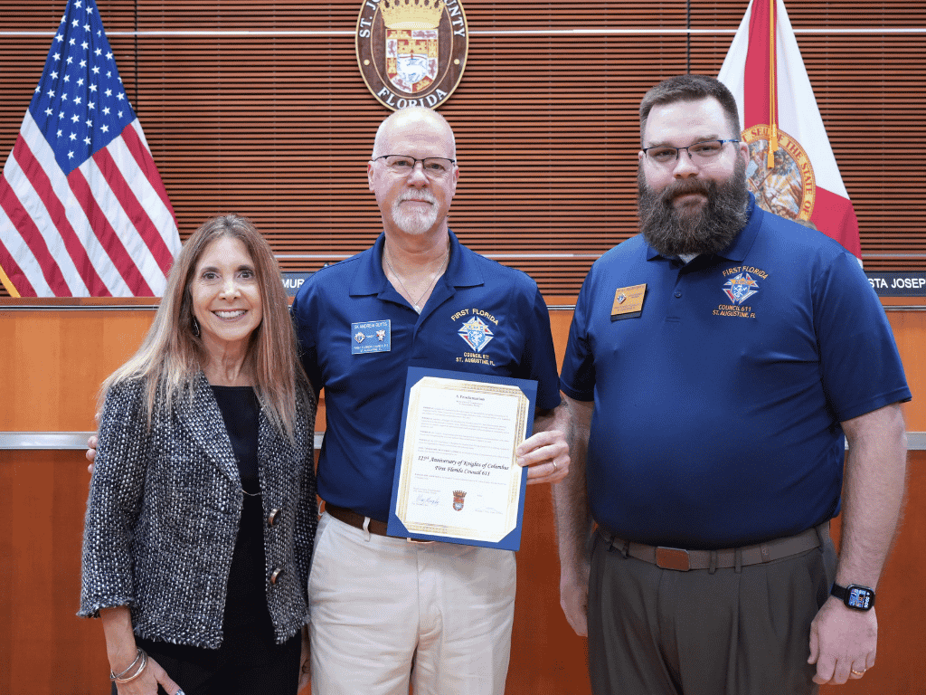 Three individuals standing together in an auditorium, one holding a certificate, with the American Flag, Florida flag, and St. Johns County Seal in the background.