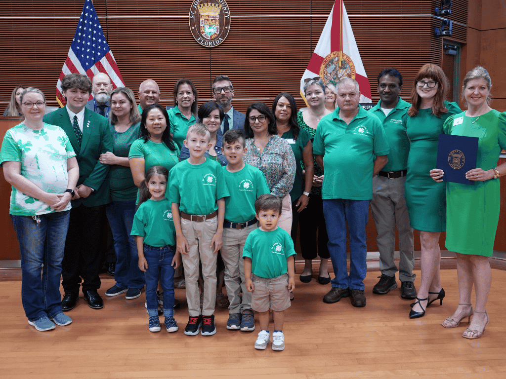 Individuals stand together, one holding a certificate, in an auditorium with the American Flag, Florida flag, and St. Johns County Seal in the background.