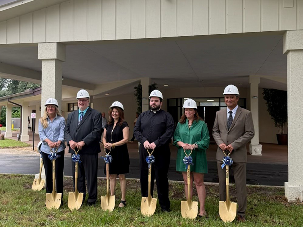Six people wearing hardhats pose for a groundbreaking photo holding gold shovels with blue ribbons.